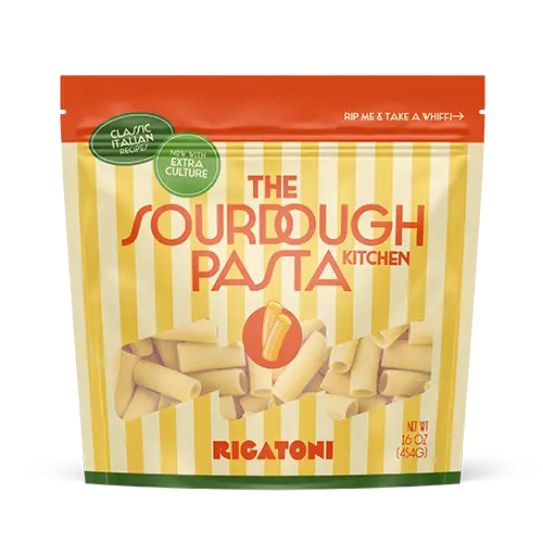 A product mockup for a pouch of The Sourdough Pasta Kitchen Rigatoni, with alternating yellow and pale yellow vertical stripes, orange text, and green info bubbles.